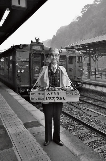 観光列車の発着時刻に合わせて人吉駅のホームに姿勢よく立つ。駅弁を満載した木箱の重量はざっと15kg。後ろの列車は特急「かわせみ・やませみ」号、右奥は「いさぶろう・しんぺい」号。