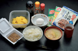 前列中央から時計回りに、ご飯、納豆、沢庵、生卵、ふりかけ（緑黄色野菜・のりたま・小魚の原料ふりかけ）、日本茶、味噌汁（キャベツ・油揚げ）。ふりかけは５～６種類を常備し、そのうち好みの２～３種が食卓に。